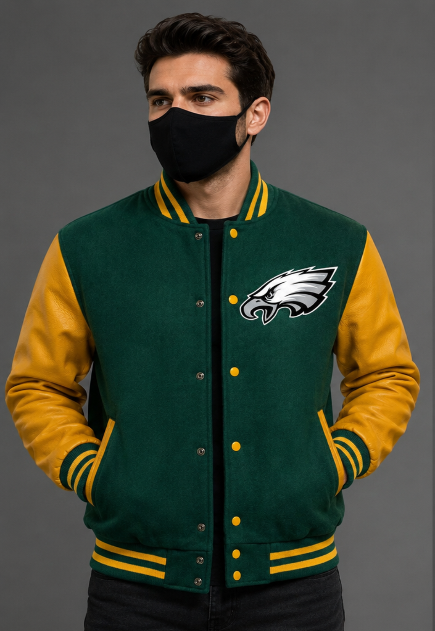 A man wearing a green and gold varsity jacket with the Philadelphia Eagles logo on the chest, paired with a black face mask, set against a solid gray studio background.