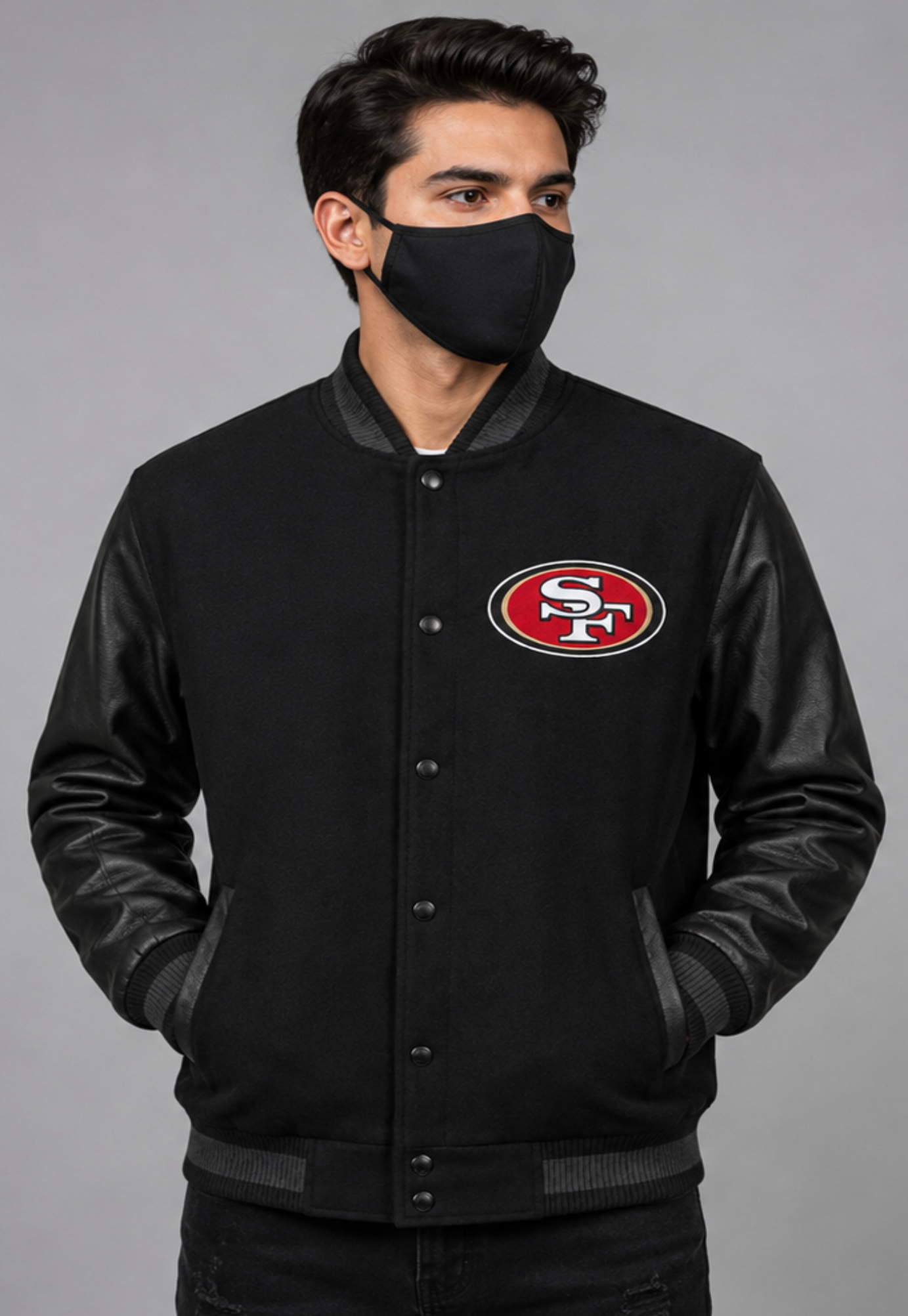 A man wearing a black varsity-style jacket with black leather sleeves and a red, white, and gold San Francisco 49ers logo on the chest, paired with a black face mask, against a solid gray studio background.