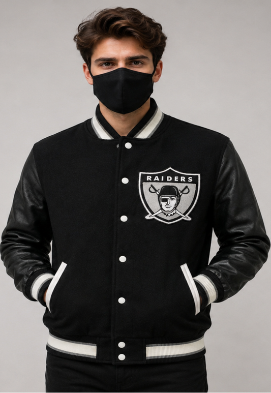 A man wearing a black Oakland Raiders varsity jacket with black faux leather sleeves, white ribbed trim, and the Raiders logo on the chest, paired with a black face mask, against a neutral gray background.