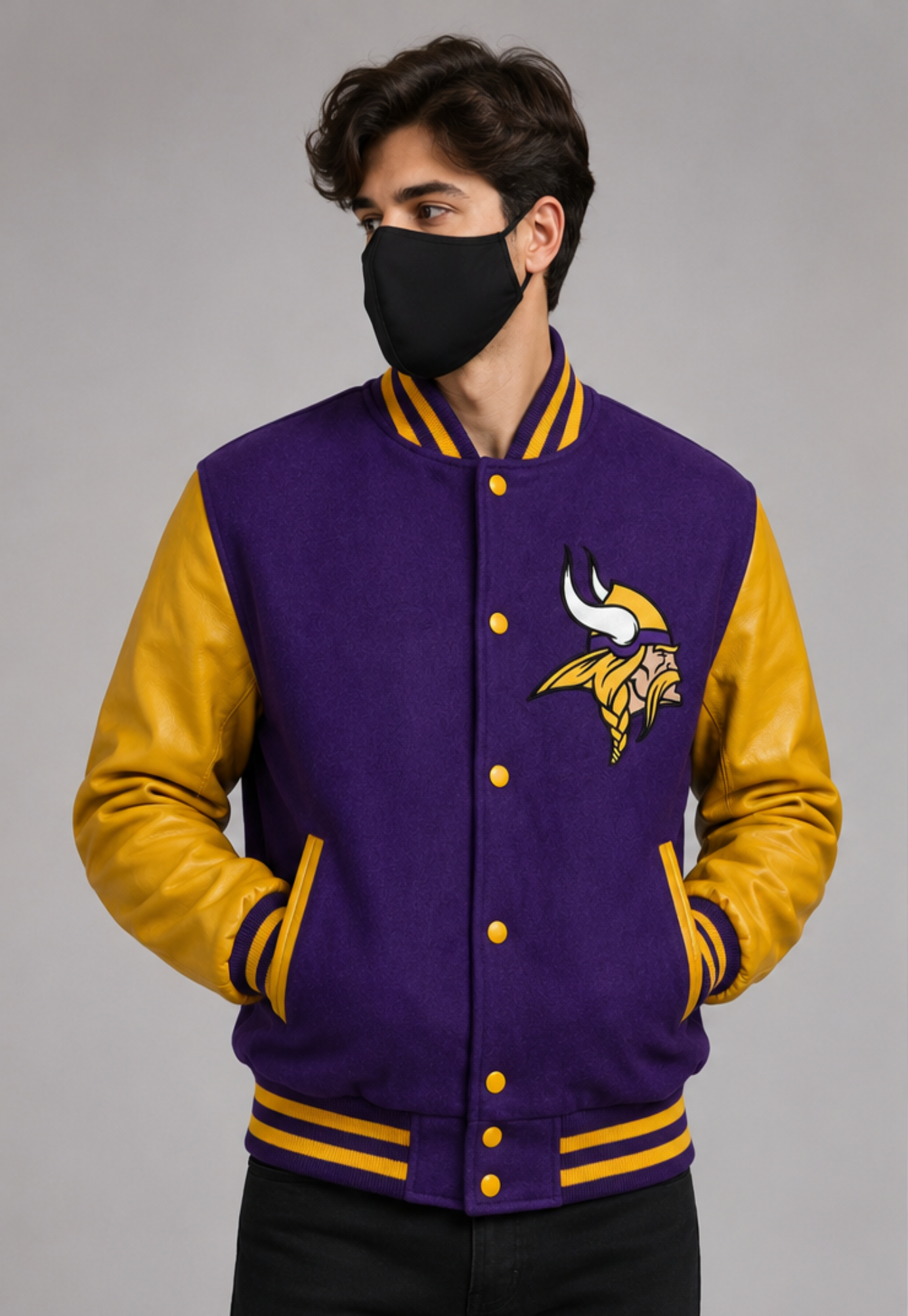 A man wearing a purple and gold Minnesota Vikings varsity jacket with a black face mask, standing against a neutral gray studio background.