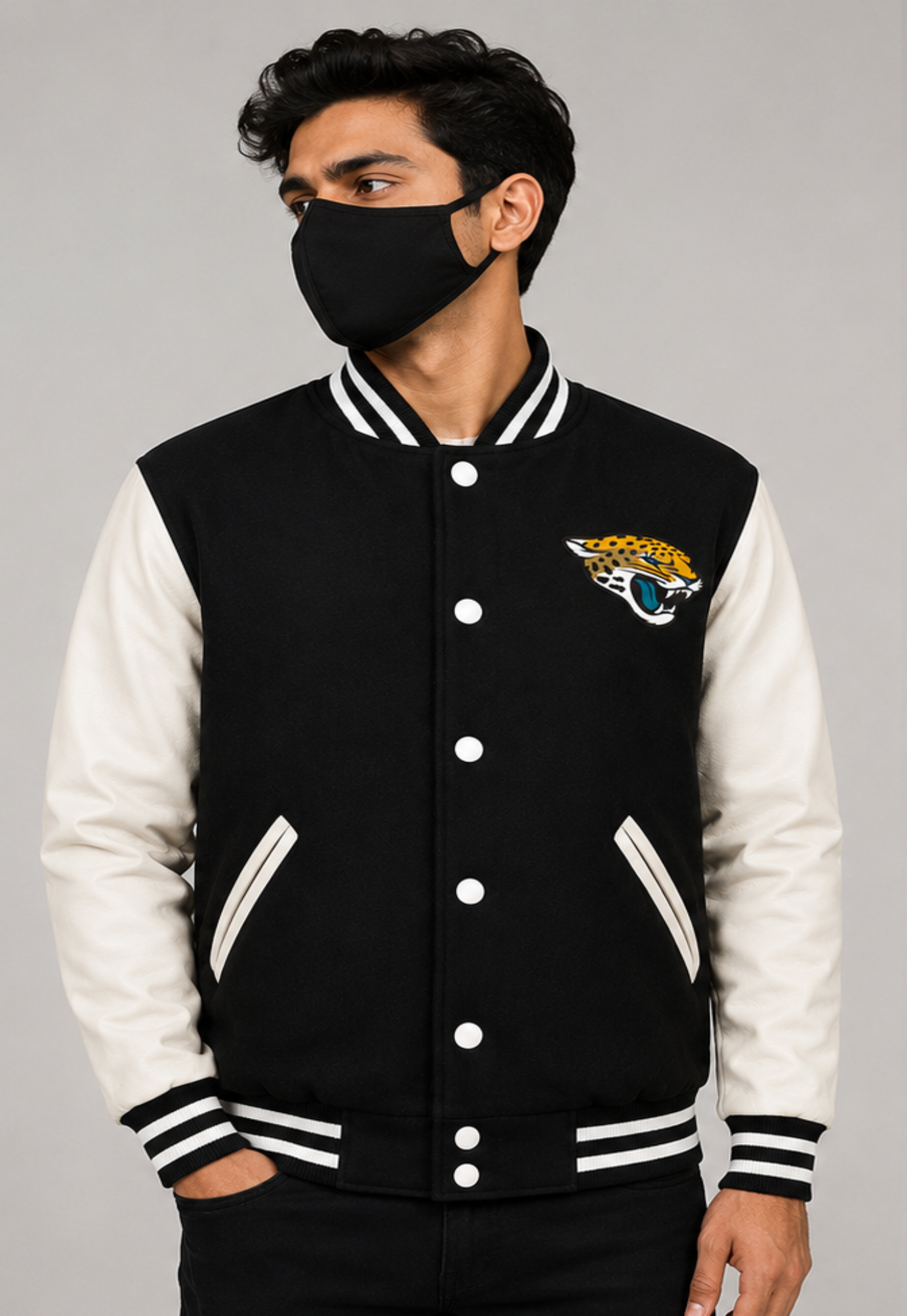 A man wearing a black and white varsity jacket with a Jacksonville Jaguars logo on the chest, paired with a black face mask, against a solid gray studio background.
