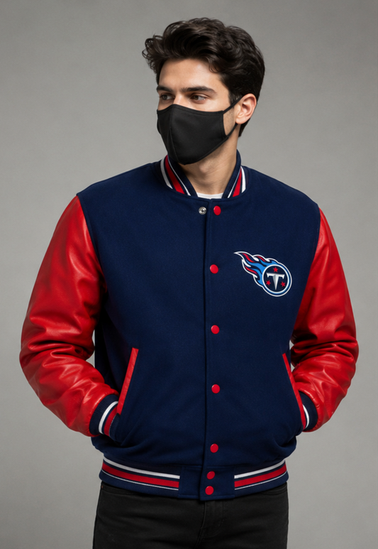 A man wearing a navy blue and red varsity jacket with the Tennessee Titans logo on the chest, paired with a black face mask, against a solid gray studio background.