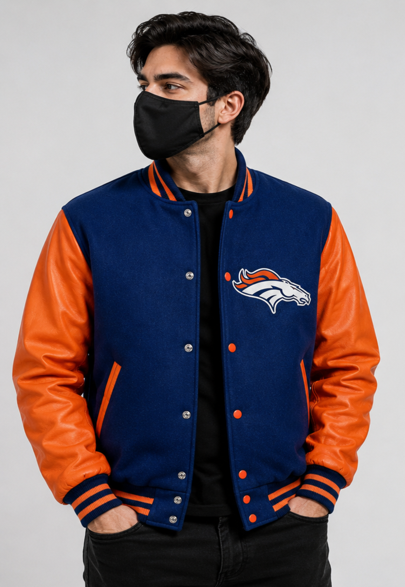 A man wearing a navy blue and orange letterman jacket with the Denver Broncos logo on the chest, paired with a black face mask and black pants, against a light gray studio background.